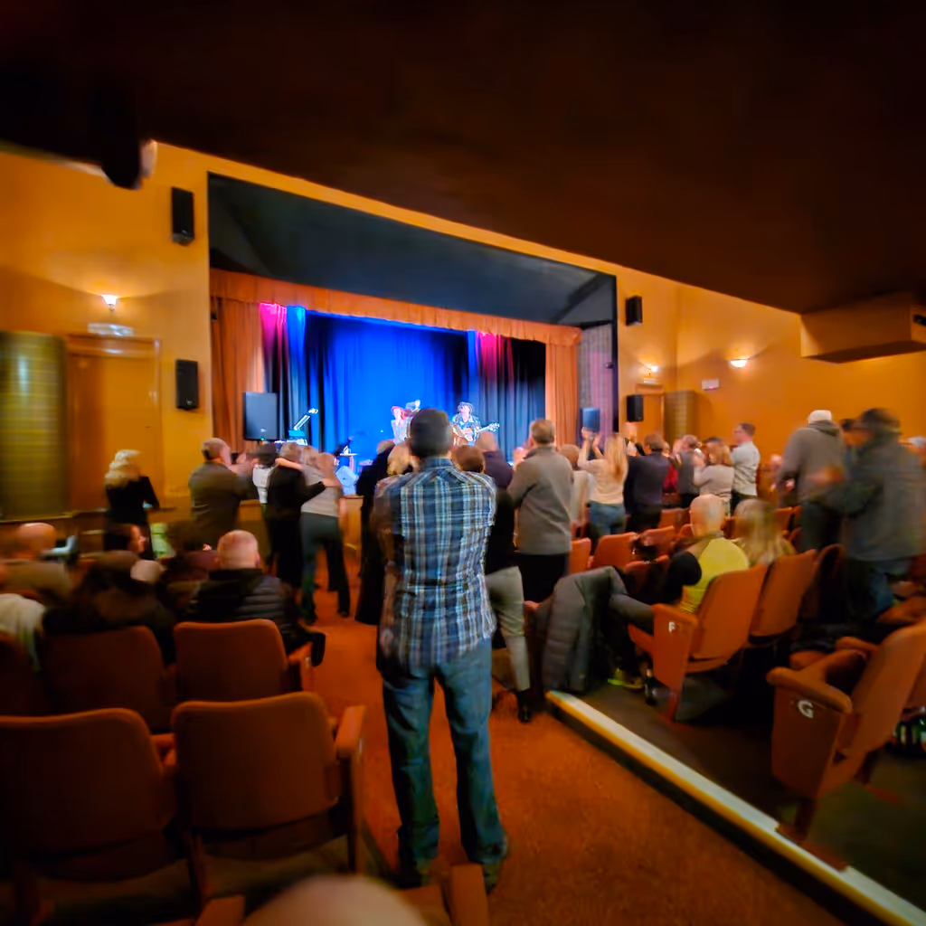 Audience clapping and standing in a small theater watching a live music performance on stage with blue and pink lighting.