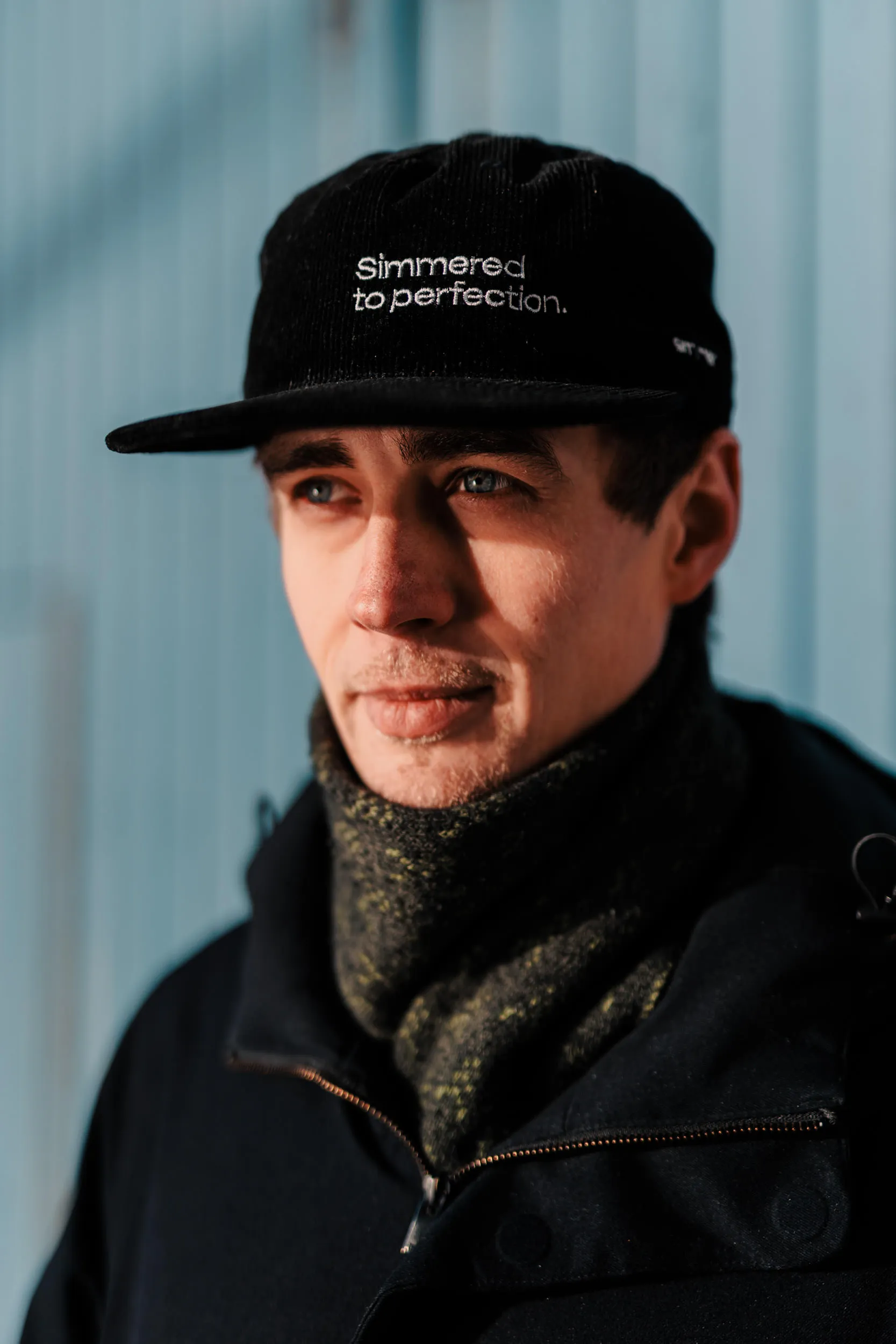 Nærbilde av mann med svart caps som har teksten 'Simmered to perfection' og en mørk vinterjakke med skjerf.