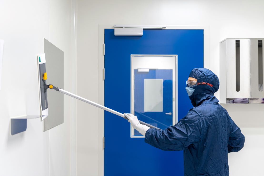 Cleanroom technician in full gowning performing routine wall cleaning, a critical component of contamination control in regulated environments. The cleanroom’s construction plays a key role in enabling effective sanitation—smooth, non-porous wall panels, flush-mounted features, and coved corners eliminate crevices where particles or microbes could accumulate.The flush doors and integrated viewing panels further enhance cleanability by minimizing surface transitions and allowing disinfectants to reach all contact points. Cleanroom architecture is deliberately engineered to support frequent, thorough cleaning procedures essential to maintaining ISO or GMP classification standards in pharmaceutical, biotech, and gene therapy facilities.