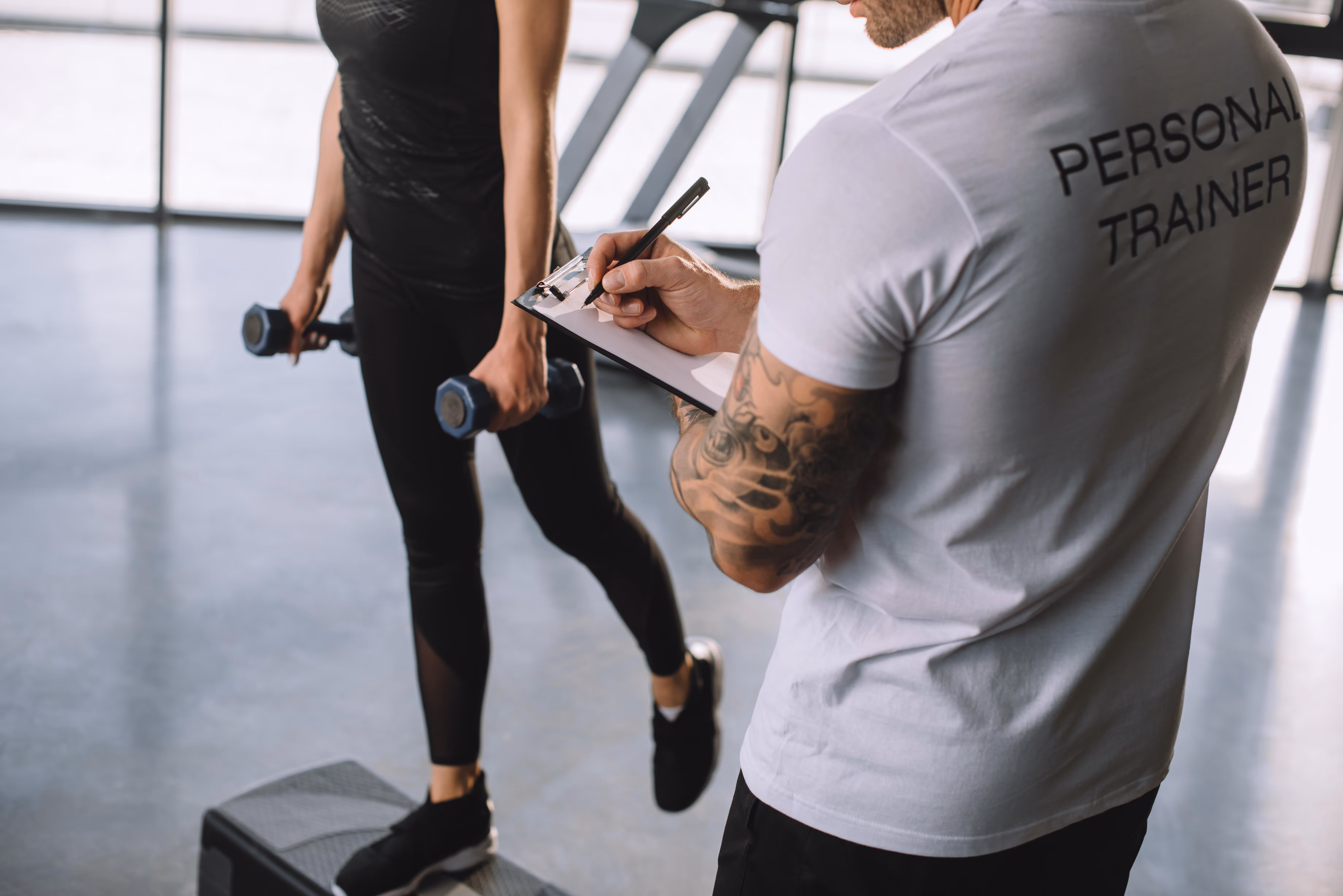 A personal trainer works one-on-one with a client, reviewing a customized fitness plan on a tablet. The trainer demonstrates proper form for a functional exercise, emphasizing strength, mobility, and personalized goals in a modern gym setting.