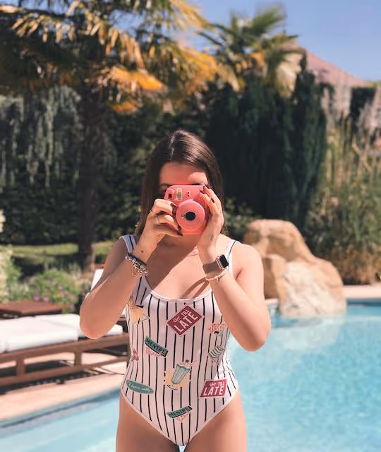 A dark haired women in a swimsuit takes a picture with a Pilot pink camera