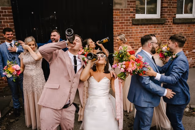 A wedding party celebrates drinking champagne from the bottle outside of a brick hotel booked on Pilot.