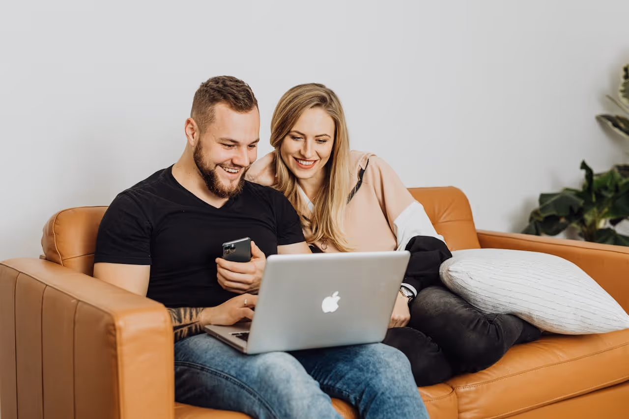 Male black shirt blue jeans sits with woman blonde hair pink shirt black pants looking at Pilot hotel room listings on silver laptop