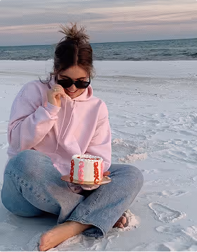 A woman in a pink hoodies eats a small birthday cake on a beach while on a trip planned on Pilot