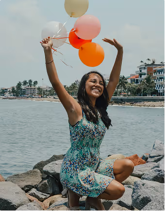 A woman in a nautical and floral dress holds 5 balloons above her head and smiles.