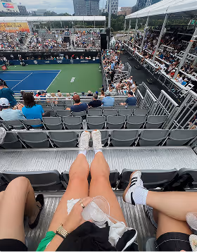 A woman sits with her legs extended in the stands at a tennis match. Her trip was planned on Pilot.