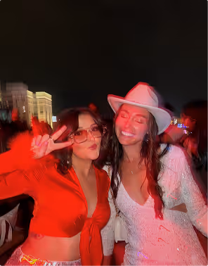 A woman in a red blouse gives a peace sign posing next to a friend in a white dress and white cowboy hat. 