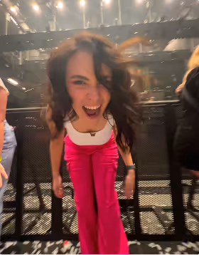 a woman in pink pants and white top screams in excitement as her trip planned on pilot got her in the front row of a concert