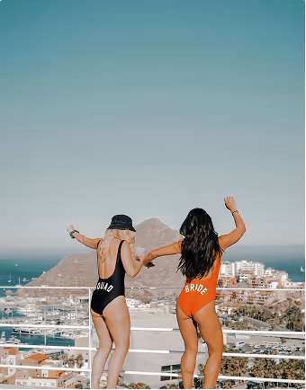 One woman wearing a black swimsuit that says squad holds the hand of a woman in a red swimsuit that says bride during a destination bachelorette party planned on Pilot. 