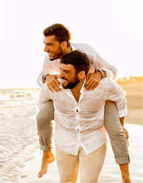 Two men in white shirts and khakis walk along the beach with one lifted onto the other's back. 