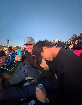 Two young men sitting outdoors in evening light, one laughing and covering his face, the other holding a green can and smiling. They are on a trip planned on Pilot. 