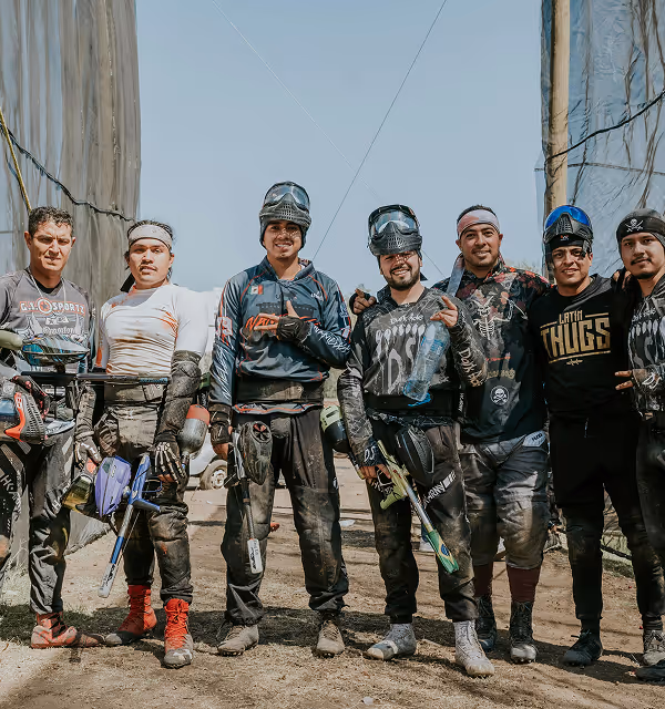 Group of seven men standing together in paintball gear holding paintball guns, smiling outdoors between netted barriers. They are on a paintball trip planned on Pilot. 