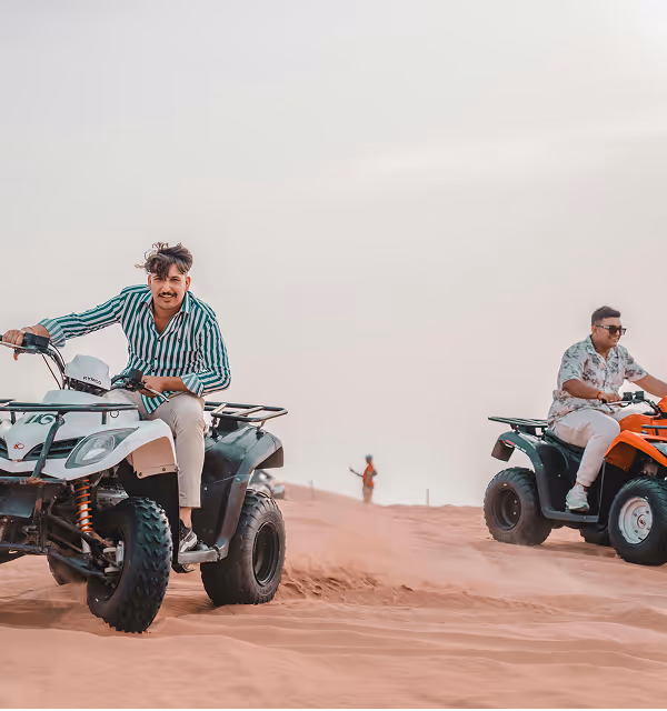 Two men riding all-terrain vehicles on sandy dunes under a clear sky during a guys trip planned on Pilot. 