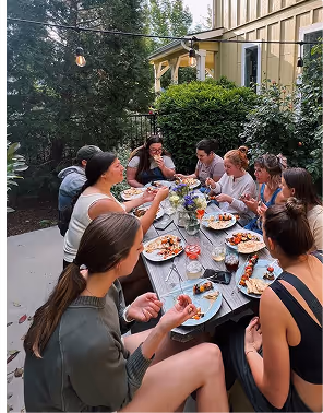 Group of people sitting around a long outdoor table enjoying a meal together in a garden setting during a foodie trip planned on Pilot.