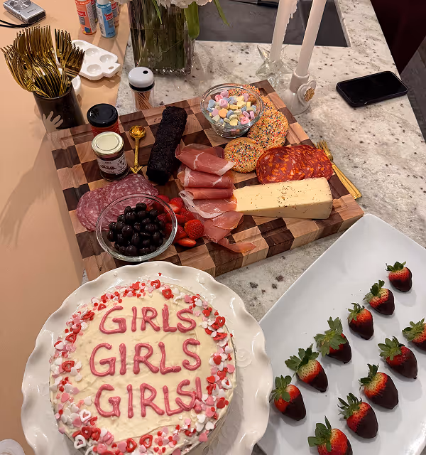 Table with a decorative cake reading 'GIRLS GIRLS GIRLS!' surrounded by heart sprinkles, a charcuterie board with assorted meats, cheese, olives, strawberries, and a bowl of heart-shaped candies, and chocolate-covered strawberries on a white plate.