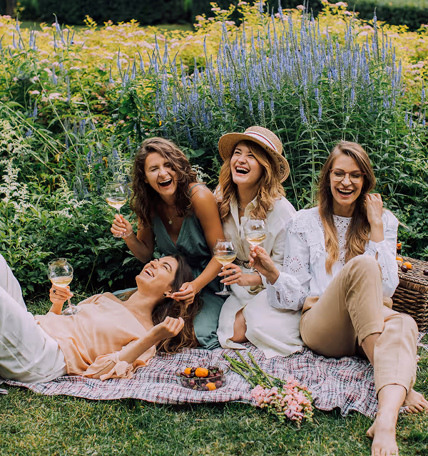 Four friends laughing and enjoying a picnic on a blanket in a garden with wine glasses and flowers during a womens holiday planned on Pilot.