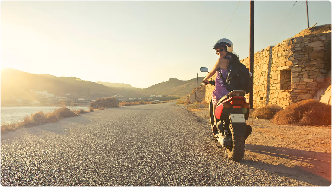 Woman wearing helmet and sunglasses riding a motorcycle on a sunlit rural road near stone walls and hills, during a backpacking trip planned on Pilot.