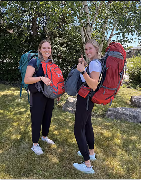 Two young women outdoors wearing large backpacks and smiling at the camera, standing on grass with trees and shrubs in the background as they plan to depart on a trip planned on Pilot. 