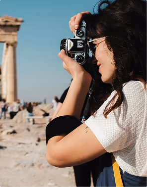 Woman with curly hair and sunglasses holding a vintage camera to her eye, taking a photo outdoors near ancient stone ruins, during a backpacking trip planned on Pilot.