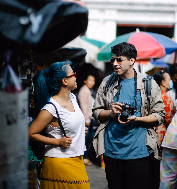 A woman with blue hair and a yellow skirt talking to a man holding a camera at an outdoor market with colorful umbrellas, during a backpacking trip planned on Pilot. 