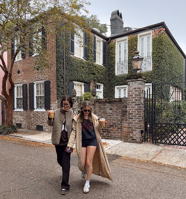 Two young women walking arm in arm holding iced coffee drinks, with ivy-covered brick house and wrought iron gate in the background, during a backpacking trip planned on Pilot. 