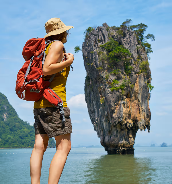 Hiker with a red backpack and hat standing near water, looking at a tall limestone rock formation in the sea, during a backpacking trip planned on Pilot.
