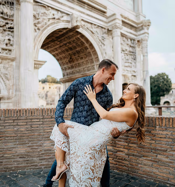 Man in patterned shirt holding and smiling at a woman in a white lace dress under an ancient stone arch, during their honeymoon planned on Pilot.