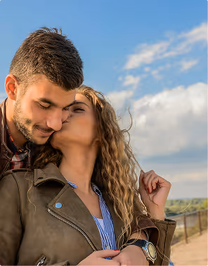 Woman with curly blonde hair kissing a man on the cheek outdoors under a partly cloudy blue sky, during their babymoon planned on Pilot.