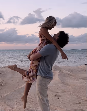 Man lifting woman on a sandy beach with the ocean and cloudy sky at sunset in the background, during their honeymoon planned on Pilot.