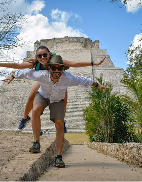 Man giving woman piggyback pose in front of an ancient stepped pyramid with blue sky, during their honeymoon planned on Pilot.