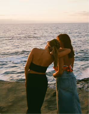 Two women standing on a rocky shore with their backs to the camera, embracing and looking out at the ocean during sunset, during their honeymoon planned on Pilot.