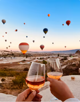 Two hands clinking glasses of rose wine with dozens of hot air balloons floating over a scenic rocky landscape at sunrise, during their anniversary tirip planned on Pilot.