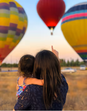 Woman holding a child and pointing at colorful hot air balloons in the sky during sunset, during a family trip planned on Pilot.
