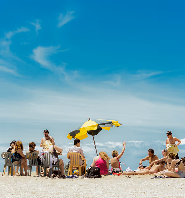 Group of people sitting and lying on the beach under a yellow and gray umbrella with a clear blue sky above, during a family trip planned on Pilot.