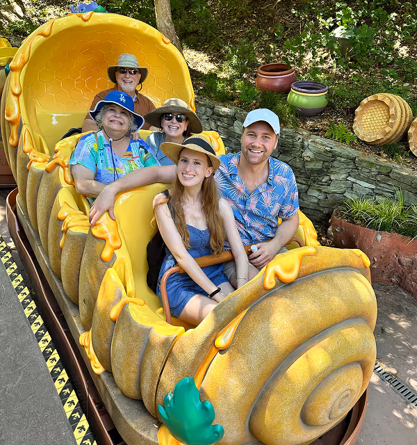 Five people wearing hats smiling while seated in a yellow snail-themed amusement park ride car, during a family trip planned on Pilot.