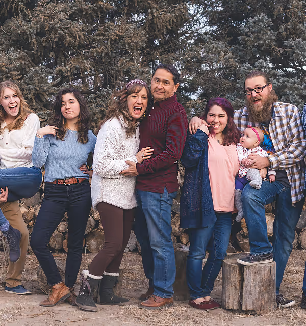 Group of seven people, including a baby, posing happily outdoors with a backdrop of pine trees and stacked wood, during a family trip planned on Pilot.