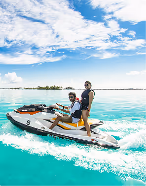 Man driving a jet ski on clear turquoise water with a woman standing behind him under a partly cloudy sky, during a beach holiday planned on Pilot. 