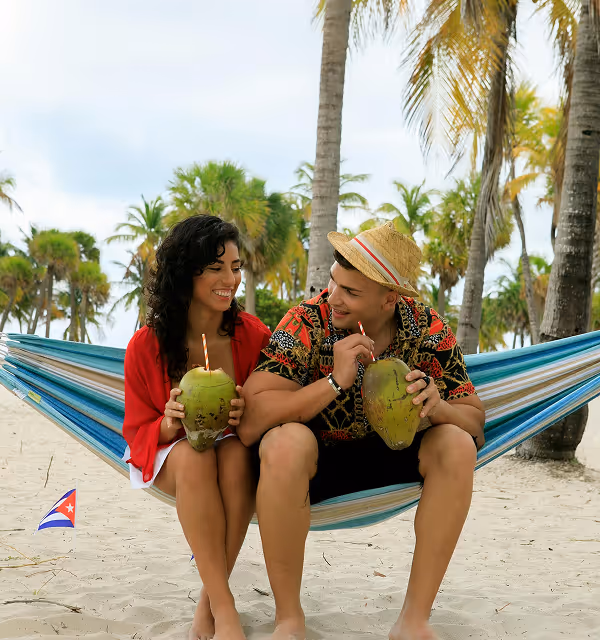 Young couple sitting on a hammock at a sandy beach, drinking from coconuts with straws, surrounded by palm trees and a small Cuban flag on the sand, during their beach holiday planned on Pilot.