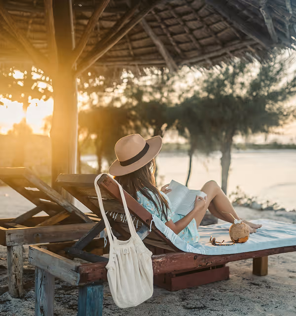 Woman wearing a hat relaxing on a wooden lounge chair under a thatched umbrella on the beach, reading a book near the water at sunset, during their beach vacation planned on Pilot. 