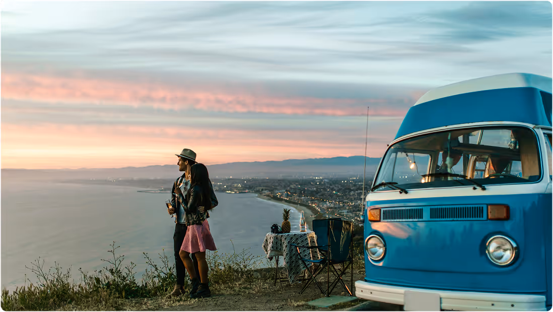 Couple standing on a hill at sunset overlooking the ocean with a blue vintage camper van and picnic setup nearby, during a road trip planned on Pilot. 