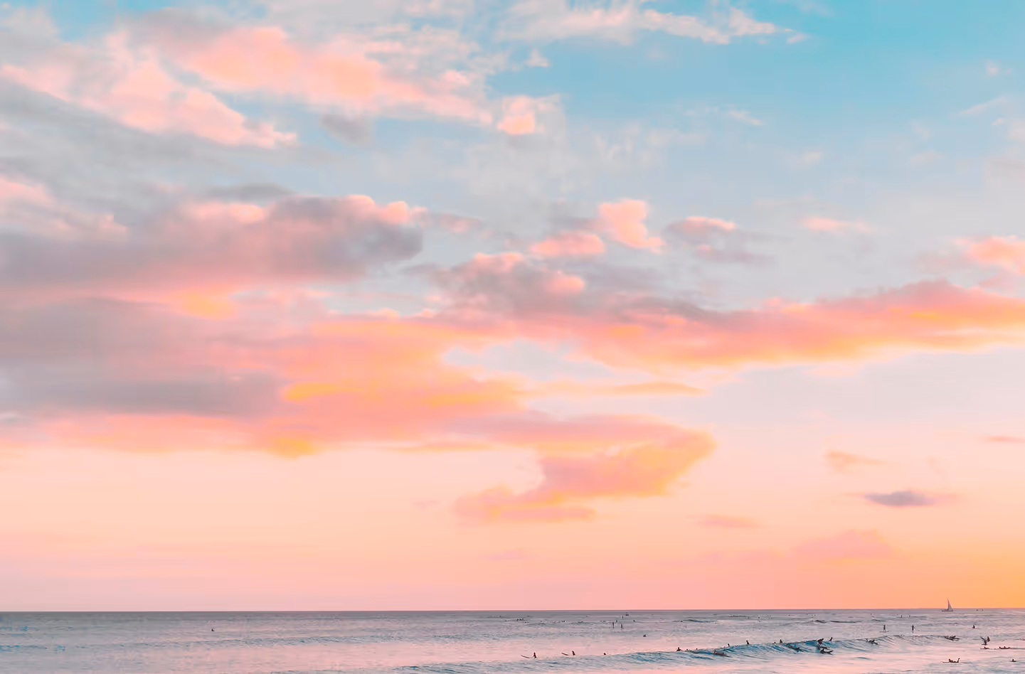 Ocean at sunset with pastel pink and blue sky and surfers in the water near the horizon.