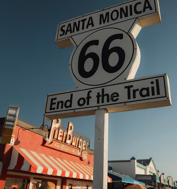 Santa Monica 66 End of the Trail road sign with Pier Burger restaurant and storefronts in the background under a clear blue sky, taken during a road trip planned on Pilot. 