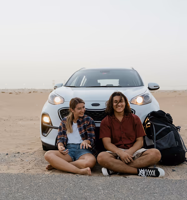 Two young people sitting barefoot on the ground in front of a white car parked in a desert, during a road trip planned on Pilot, with a large backpack beside them.