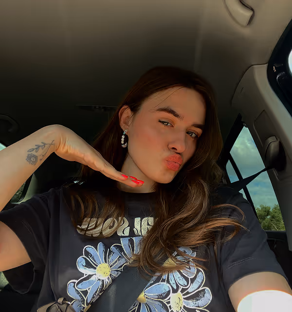 Young woman during a road trip planned on Pilot with long brown hair taking a selfie in a car, puckering lips and posing with a hand under her chin, wearing a black t-shirt with a daisy print and a seatbelt.