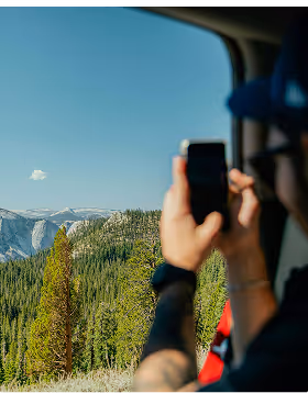 Person inside a vehicle during a road trip planned on Pilot taking a photo of a scenic mountain landscape with pine trees under a clear blue sky.