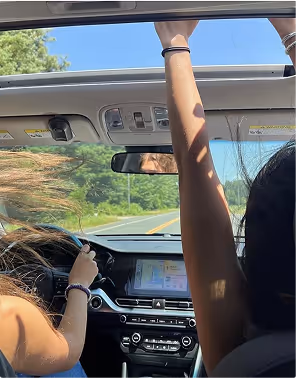 Two people in a car on a sunny day, during a road trip planned on Pilot, one driving with hair blowing, the other raising an arm through the open sunroof.