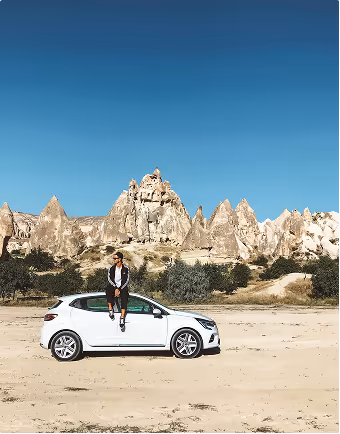 Person sitting on the roof of a white car during a road trip planned on Pilot parked on a sandy area with rocky hills and a clear blue sky in the background.