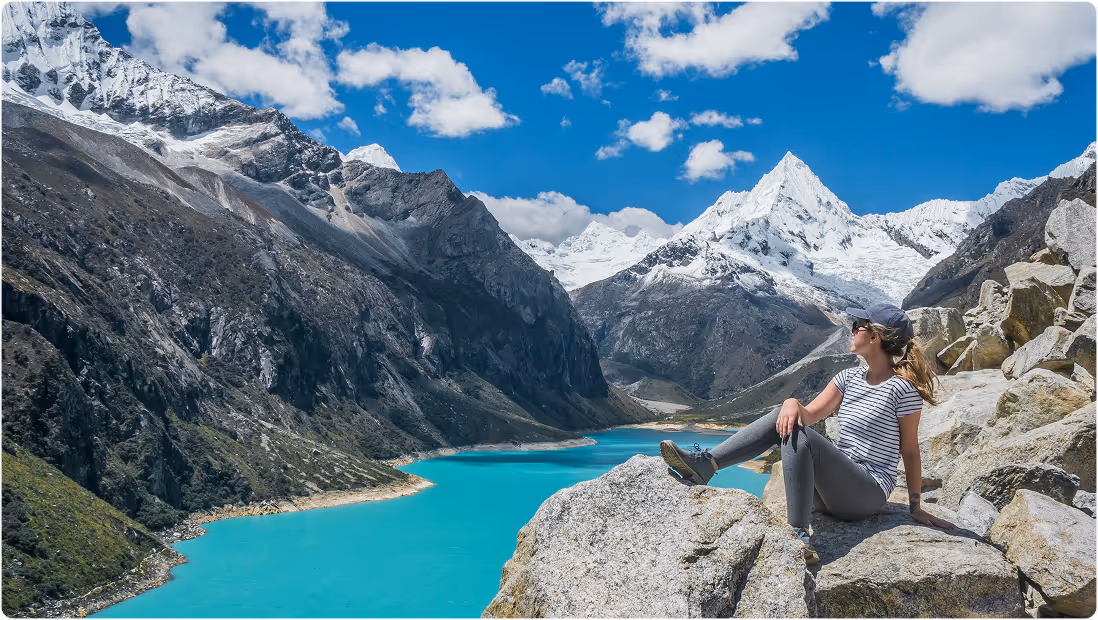 Woman sitting on rocks overlooking a turquoise lake surrounded by snow-capped mountains under a blue sky with scattered clouds, during her trip planned on Pilot.