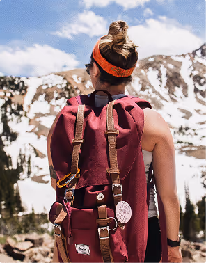 Person with a maroon backpack and orange headband looking at snow-covered mountains under a partly cloudy sky, during her travels planned on Pilot.