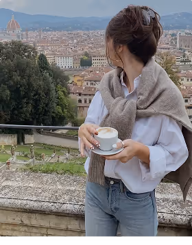 Woman holding a cup of coffee overlooking a cityscape with historic buildings and a cathedral dome in the background, during her travels planned on Pilot.